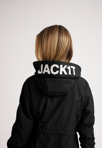 Giacca nera con cappuccio, con un logo bianco "JACK1T" sul colletto. Il tessuto appare liscio e leggero.