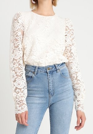 Top di pizzo in avorio con maniche lunghe a sbuffo, caratterizzato da motivi floreali. Abbinato a jeans in denim blu a vita alta con una leggera elasticità.