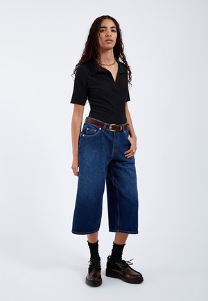 Donna dai capelli ricci che indossa una maglietta nera a maniche corte, jeans blu larghi corti, cintura marrone, calzini neri e scarpe nere stringate.