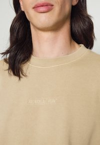Sudadera beige con cuello redondo, puños y dobladillo acanalados. Presenta un estampado en relieve de la marca "REVOLUTION" en el pecho. Textura suave, diseño casual.