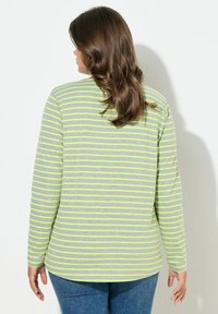 Žena s dlouhými hnědými vlnitými vlasy, která má na sobě šedé tričko s dlouhým rukávem s neonově zelenými pruhy a modré džíny, čelem ke bílému zdi.