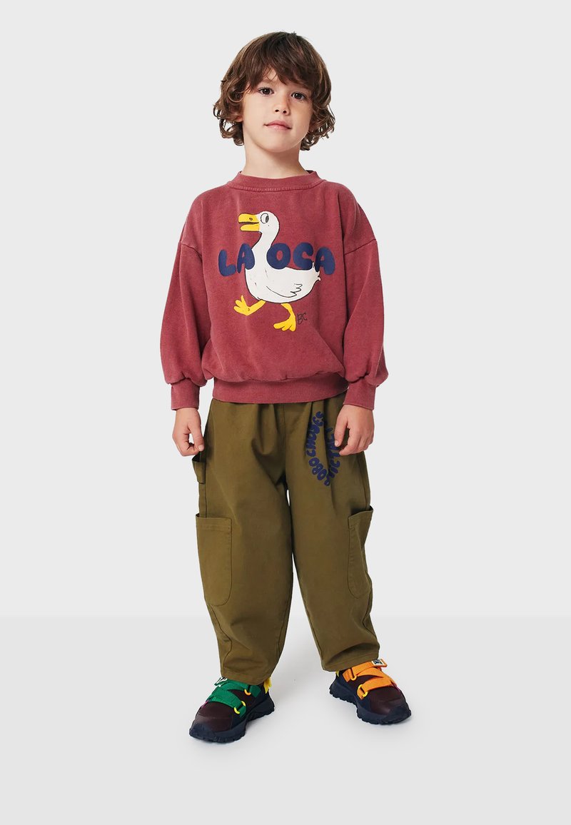 Bambino con un maglione bordeaux con la scritta "LA OCA" e una grafica di un'anatra, abbinato a pantaloni cargo verdi e scarpe sportive multicolori.