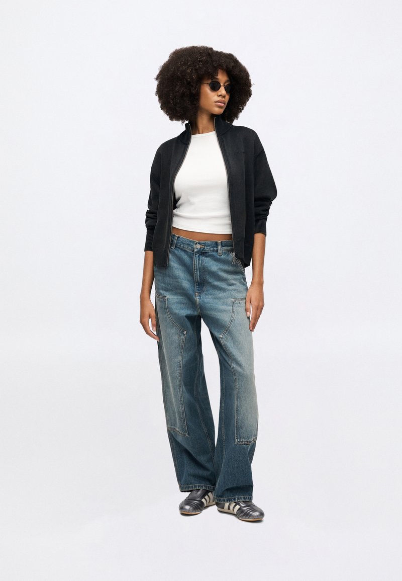 Black zip-up bomber jacket over a white crop top, paired with light blue wide-leg jeans. Model wears round black sunglasses and silver sneakers.