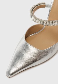 Silver metallic high-heeled shoe with a pointed toe and textured finish. Features a decorative rhinestone ankle strap.