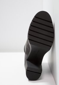 Semelle noire d'une botte en daim gris placée à la verticale, montrant le motif rainuré de la semelle et un talon carré sur fond blanc.