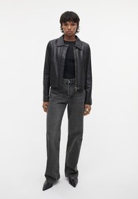 Schwarze Lederjacke mit Kragen über einem schwarzen Oberteil, kombiniert mit hoch taillierten, verblassten grauen weit geschnittenen Jeans und spitzen schwarzen Schuhen.