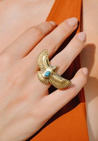 Bague en or en forme d'oiseau avec des ailes texturées et une pierre turquoise au centre, portée sur une main posée sur un fond en tissu orange.
