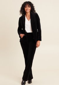 Blazer in velluto nero con rever a punta, tasca singola, abbinato a pantaloni coordinati. Camicia bianca a scollo a V sotto. Texture liscia.