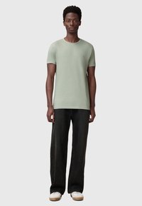 Camiseta de algodón verde claro con cuello redondo, mangas cortas y un pequeño detalle de logo. Combinada con pantalones de mezclilla anchos negros y zapatillas blancas.