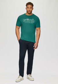 Groene T-shirt met korte mouwen en een wit tekstontwerp met "Hooked On Scouting Fly Fishing Equip." Gecombineerd met donkere broek en lichte sneakers.