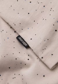 Světlé béžové bavlněné plátno s malými černými a modrými skvrnami. Obsahuje přišité štítek s textem "simple wear". Měkká textura, neformální design.