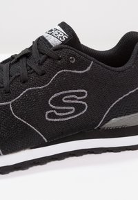 Czarne siatkowe buty sportowe z szarymi akcentami, z teksturowaną cholewką, sznurowadłami i białą podeszwą środkową. Logo jest wyraźnie widoczne z boku.