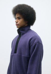 Violets flīsa džemperis ar melnu ar rāvējslēdzēju pusi apkakli, ar teksturētu virsmu un sānu kabatu, parādīts no sāniem.