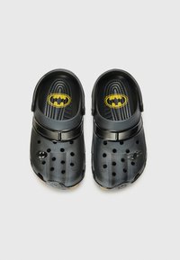 Black clogs featuring a Batman logo in yellow, with ventilation holes and a textured, smooth surface. Lightweight and casual design.