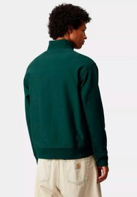 Sudadera verde de cuello alto hecha de tela suave. Cuenta con puños y dobladillo acanalados. Se lleva con pantalones de color claro que muestran el logo de la marca en el bolsillo.