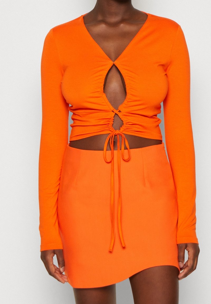 Orange långärmad topp med djup V-ringning och nyckelhålssömm, kombinerad med en matchande orange minikjol som har en böjd nederkant.