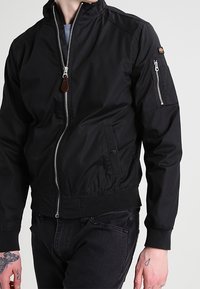 Czarna kurtka bomberka wykonana z lekkiej tkaniny, z wysokim kołnierzem, przednim zamkiem błyskawicznym oraz zamykana kieszenią na lewym rękawie. Elastyczne mankiety i dół kurtki.