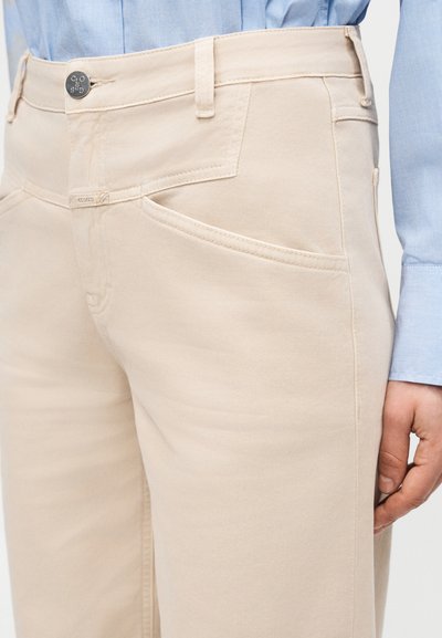 Pantalon beige avec coutures courbes sur les poches avant, passants pour ceinture, bouton en métal gravé "CLOSED", porté avec une manche de chemise bleu clair visible.