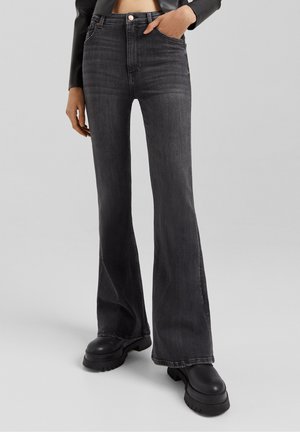 Personne portant un jean évasé noir délavé taille haute avec des bottes noires épaisses et une veste courte en cuir noire, la main dans la poche.