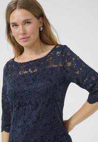 Top in pizzo blu navy con maniche a tre quarti, motivo floreale e scollatura a barchetta. Tessuto morbido con dettagli trasparenti e bordi ondulati.