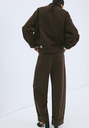 Personne aux cheveux tressés portant une veste ample brun foncé et un pantalon large assorti, debout, les mains sur les hanches, vue de dos.