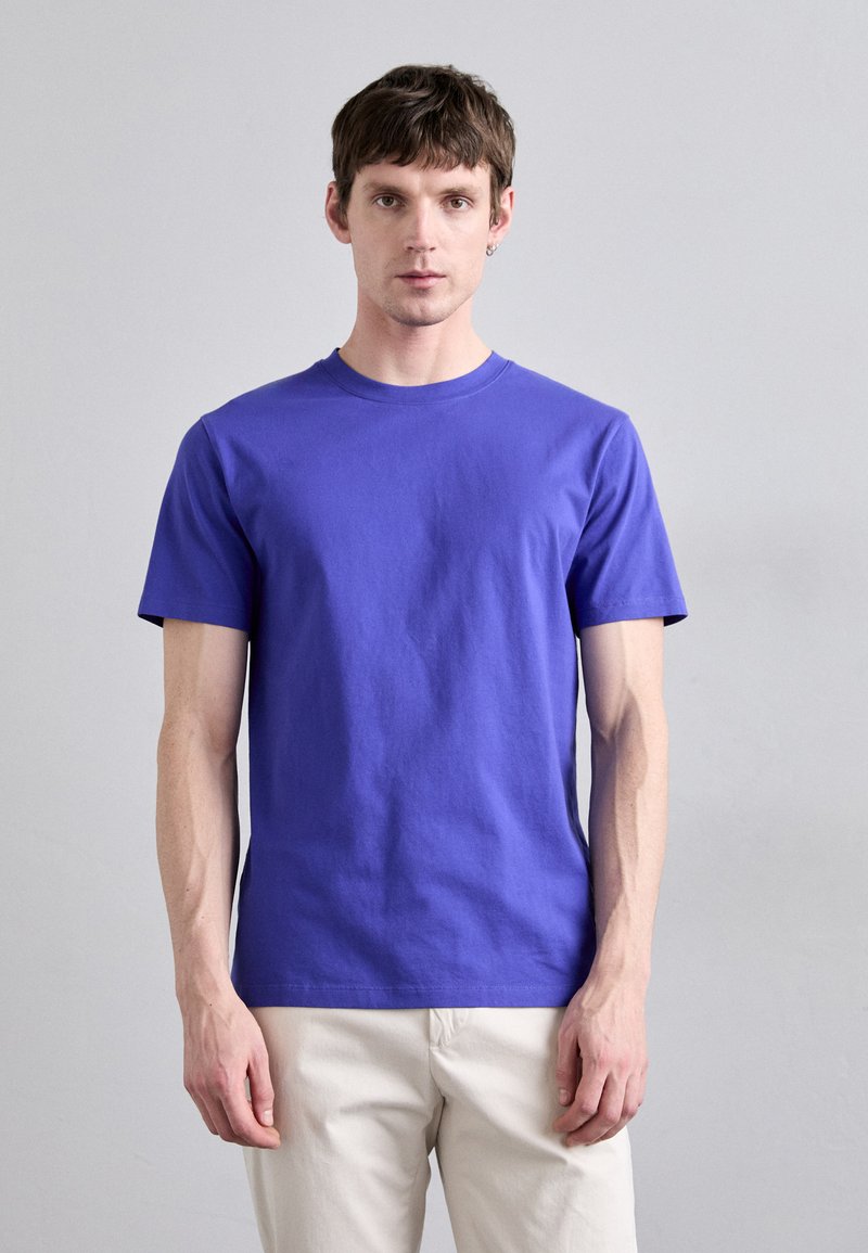 T-shirt à manches courtes, col rond, en tissu de coton violet uni. Texture lisse avec une coupe régulière. Porté avec un pantalon de couleur claire.