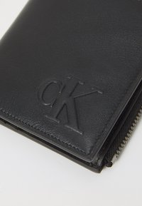 Svart skinnlommebok med embossert "CK"-logo, glidelåslukking, teksturert overflate og ren søm langs kantene.