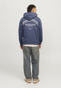 Sudadera azul marino con un gran estampado de texto blanco en la parte posterior. Presenta un bolsillo tipo canguro, capucha ajustable y puños acanalados. Combinada con jeans claros.