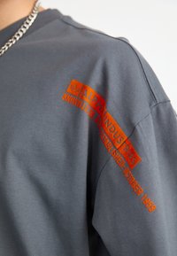 T-shirt en coton gris avec texte de logo orange sur la manche gauche. Coupe décontractée avec un col rond et des manches courtes.