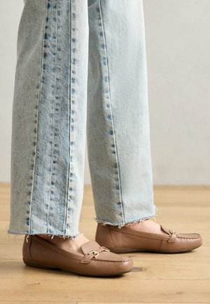 Bruine leren loafers met een gestructureerde afwerking, voorzien van een decoratieve band en gouden beslag; gedragen met lichtblauwe spijkerbroek met rafels op een houten vloer.