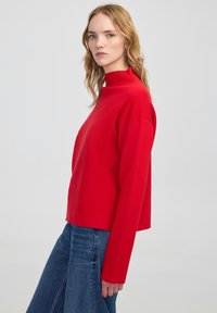 Red long-sleeve sweater with a high neck, cropped design, soft texture, and a relaxed fit. Paired with blue flared jeans.