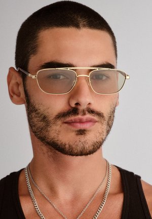 Mężczyzna z krótkimi włosami i brodą, noszący złote okulary przeciwsłoneczne, czarny tank top i dwa srebrne łańcuszki, neutralne tło.