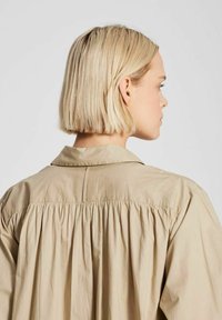 Chemise beige à boutons avec col, présentant un tissu froncé au dos pour ajouter de la texture et une coupe décontractée.