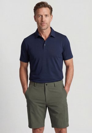 Homme portant un polo bleu marine et un short cargo vert olive, debout, les mains dans les poches, sur un fond clair uni.
