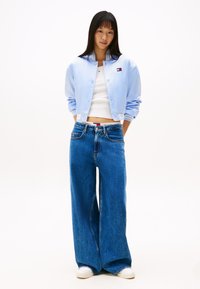 Ungdommelig kvinne iført lyseblå bomberjakke, hvit kort topp, brede blåjeans og hvite joggesko mot hvit bakgrunn.