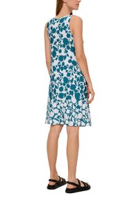 Ein ärmelloses, knielanges Kleid mit floralem Muster in Blau auf leicht blauem Hintergrund, kombiniert mit schwarzen flachen Sandalen.