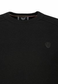 Schwarzer Strickpulli mit gerippter Textur und rundem Halsausschnitt. Er verfügt über ein kleines erhabenes Wappen-Logo auf der Brust.