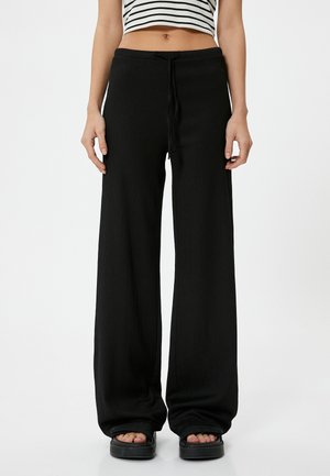 Schwarze weit geschnittene Hose mit gerippter Textur, elastischem Kordelzug und fließender Silhouette, kombiniert mit klobigen, offenen Schuhen.