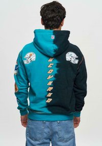 Sweat-shirt à capuche avec un design dégradé, fusionnant le teal et le noir. Il présente des logos de la NFL, le texte "COVERED" et des accents contrastants sur les manches.