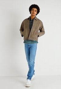 Jaqueta de denim castanha sobre uma camisa às riscas verdes e brancas, combinada com umas calças de ganga azul claro e ténis brancos. Design casual e descontraído.