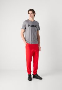 Grijze t-shirt met groot zwart "HUGO" logo, gecombineerd met rode joggerbroek. Model draagt zwarte sneakers. Eenvoudig, modern outfitontwerp.