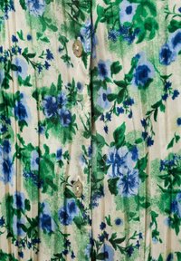 Bloemige stof met groene en blauwe bloemen op een crèmekleurige achtergrond, met een knoopsluiting en een licht gestructureerd oppervlak.