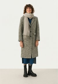 Manteau gris texturé avec de grands boutons, associé à une écharpe beige clair. Porté par-dessus une robe en denim et des bottes sombres à semelles épaisses.