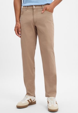 Mann trägt beige gerade geschnittene Hose mit Fronttaschen und weiße Sneakers mit braunen Streifen, steht vor weißem Hintergrund.