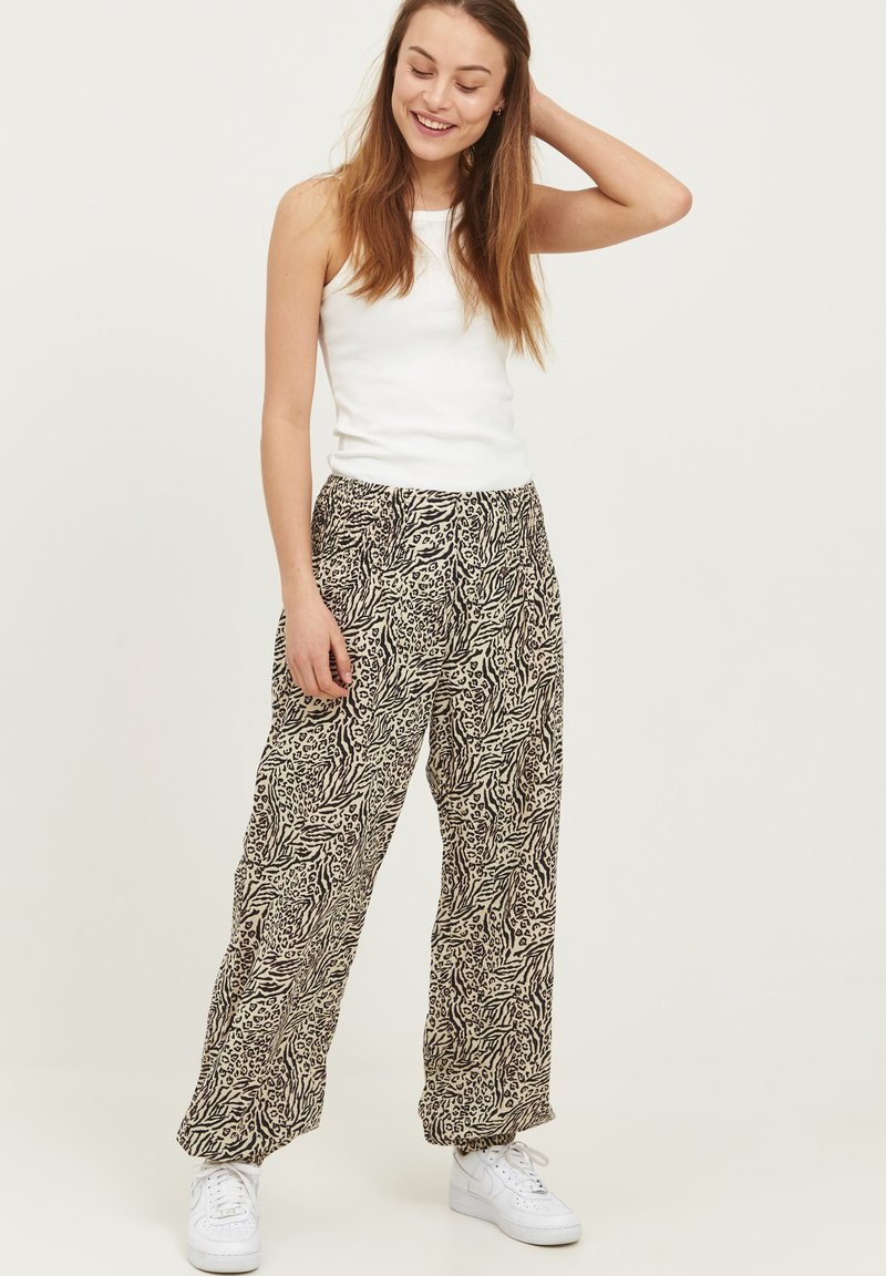 Losse broek met een zwart-beige dierenprint, gemaakt van lichtgewicht materiaal, gecombineerd met een wit tanktop en witte sneakers.