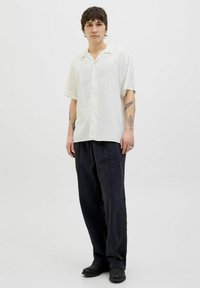 Kortærmet, off-white skjorte med krave og knapper, parret med mørke, løstsiddende bukser. Personen bærer sorte sko og viser tatoveringer.