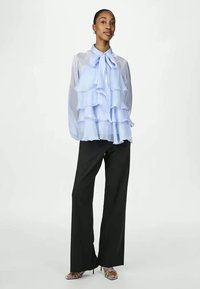 Világoskék fodros blúz nyakkendővel a nyaknál, fekete széles szárú nadrággal és pántos magassarkúval, semleges háttér előtt.