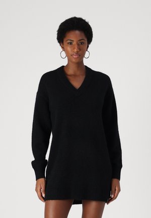 Robe pull noire oversize à col en V, confectionnée en tissu doux et texturé, avec des manches longues et des poignets et bas côtelés, présentant une coupe décontractée.