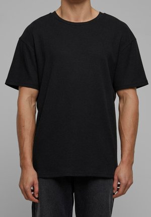 Mann trägt ein einfaches schwarzes T-Shirt mit kurzen Ärmeln und Rundhalsausschnitt sowie dunkle Hose vor einem neutral grauen Hintergrund.