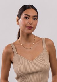Goldene Kettenhalskette mit unregelmäßigen weißen Perlen. Getragen mit einem beigen gerippten Tanktop, das ein V-Ausschnitt-Design zeigt.